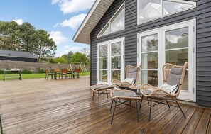 Une terrasse avec des chaises et une table en osier, une balançoire et un barbecue à l’arrière-plan.