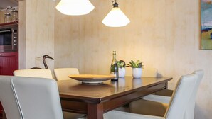 A dining area with a wooden table, white chairs, two pendant lights, a bottle of champagne, and two potted plants.