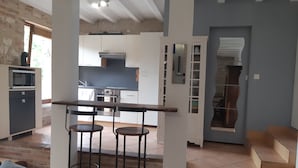 Une cuisine moderne avec des armoires blanches, un four encastré et un four à micro-ondes. On y trouve un coin repas avec une table en bois et deux tabourets. La pièce est dotée d’un mur en briques et d’une grande fenêtre.