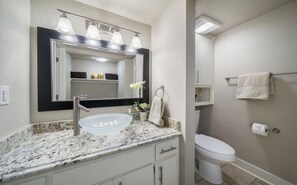 A bathroom with a granite countertop, a white sink, a mirror with lights, a toilet, and a towel rack.