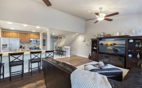 A spacious living area with a large flat-screen TV, a leather couch, and a dining area with a kitchen in the background.