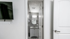 Un couloir étroit mène à une salle de bain dotée d’un plancher en bois, d’un miroir rond et d’une porte blanche.