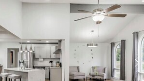 Une cuisine moderne dotée d’un îlot central, d’un coin repas avec des tabourets de bar, d’un espace détente avec un canapé et une table basse, et d’une grande fenêtre agrémentée de rideaux.