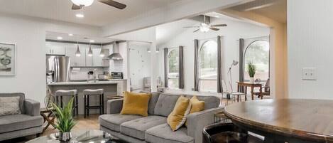 A spacious living room with a gray sofa, a coffee table, and a dining area with a wooden table and chairs. There is a kitchen in the background with white cabinets and stainless steel appliances.