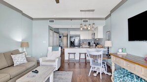 Una sala moderna con un sofá, una mesa de centro y un televisor. Cuenta con una zona de cocina con refrigerador, microondas y taburetes de bar.
