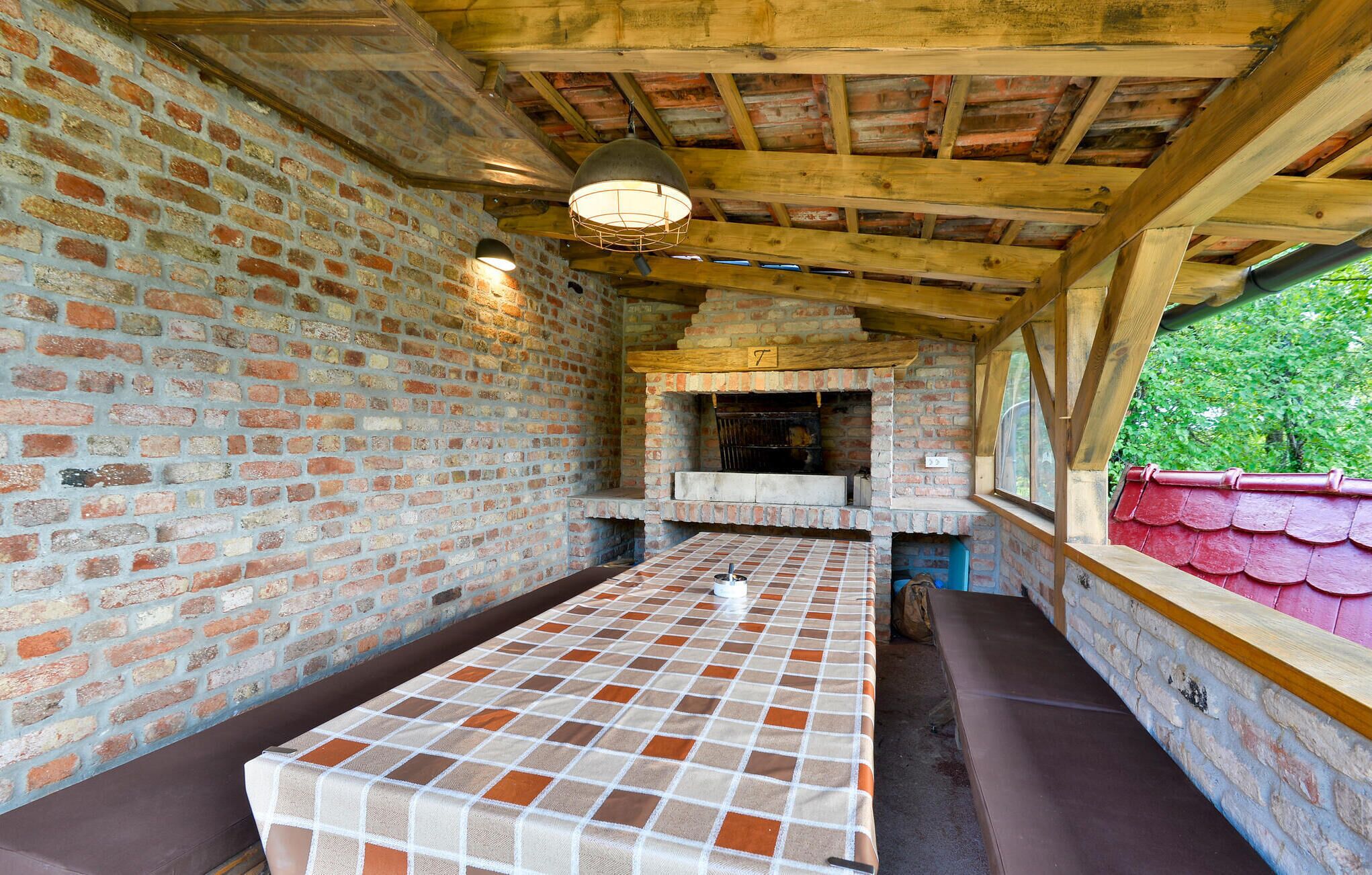 A rustic brick interior with a tiled table and benches, a brick fireplace, and a wooden ceiling.