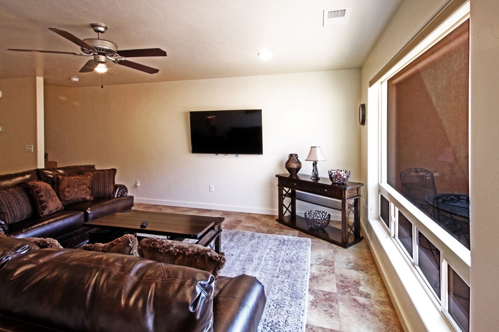 A living room with a leather sofa, a coffee table, a TV mounted on the wall, a ceiling fan, and a large window.