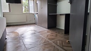 Une pièce dotée d’un parquet à motifs en chevrons, d’une grande fenêtre avec des stores et d’un radiateur.
