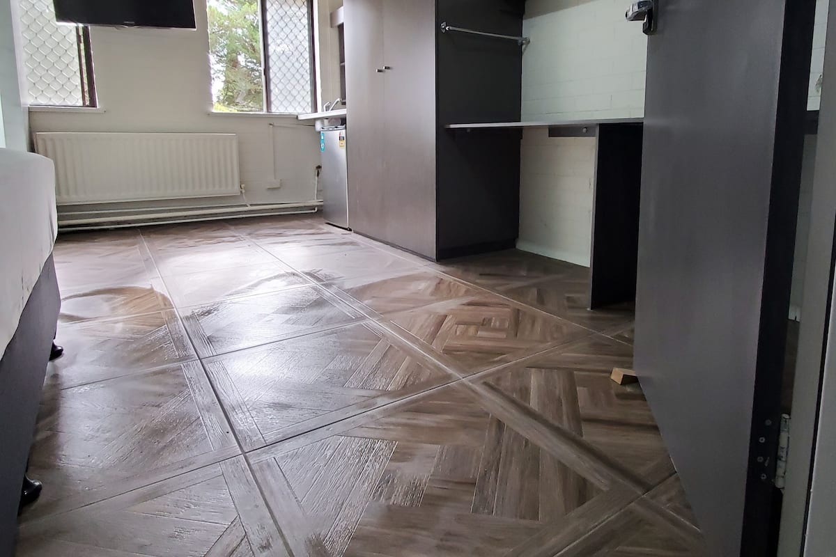 A room with a herringbone-patterned floor, a large window with blinds, and a radiator.