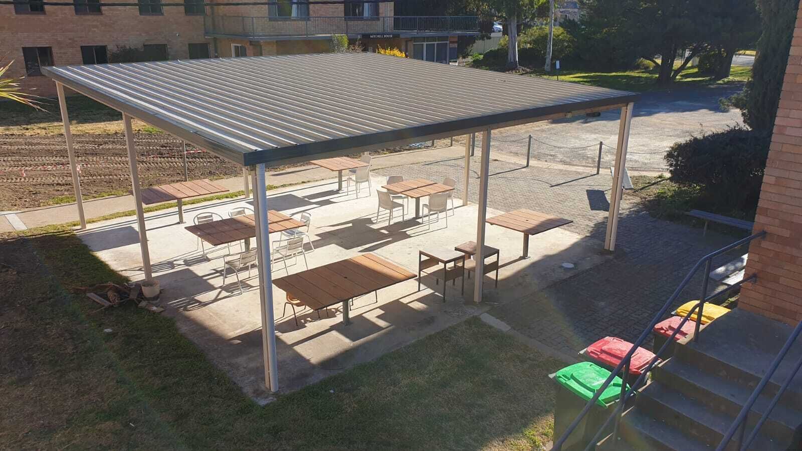 An outdoor patio area with tables and chairs, a covered seating area, and a brick building in the background.