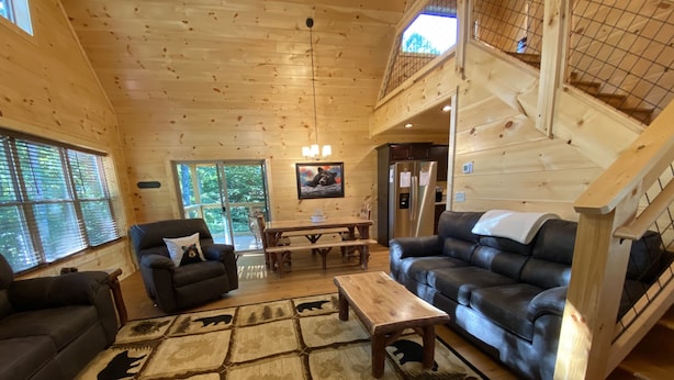 Ein gemütliches Blockhaus-Interieur mit Holzbalkendecke, einem Essbereich, einem Wohnzimmer mit Sofa und Couchtisch sowie einer Treppe.
