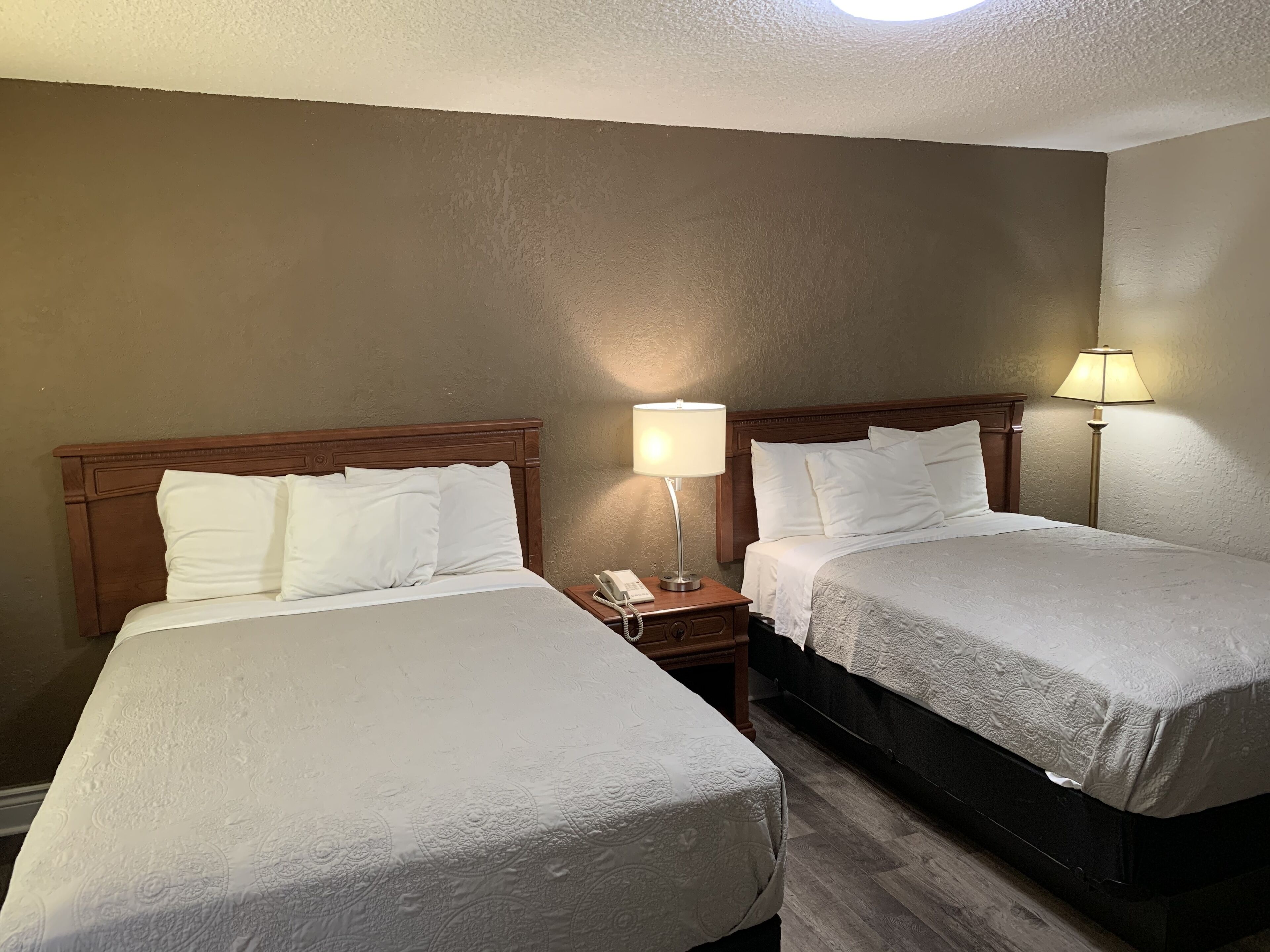 A hotel room with two beds, each with white bedding and a wooden headboard. There is a nightstand with a lamp and a phone between the beds.