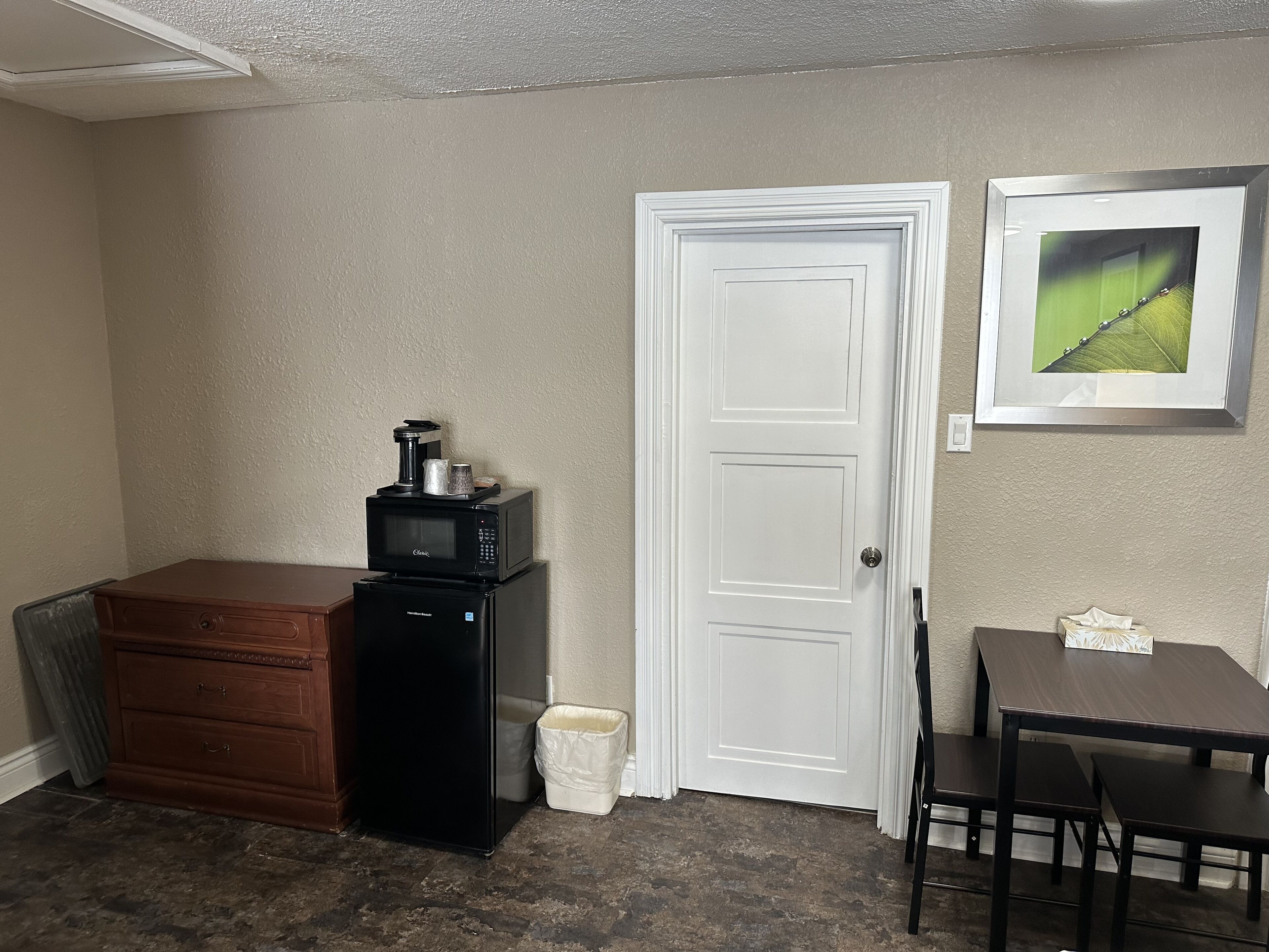 A compact kitchen with a refrigerator, microwave, and coffee maker on a counter, a wooden cabinet, a trash bin, a white door, and a small table with chairs.