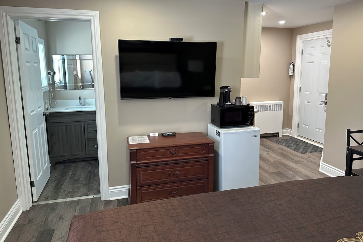 A compact hotel room with a kitchenette, a flat-screen TV, a small refrigerator, and a wooden dresser.