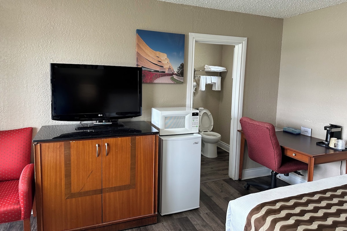 A hotel room with a television, a microwave, a small refrigerator, a desk with a chair, a bed, and a bathroom with a toilet and towel rack.