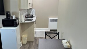 A compact kitchen with a microwave, coffee maker, and a small refrigerator. There is a dining table with chairs and a trash bin. - Champlain Motor Inn (Pembroke)