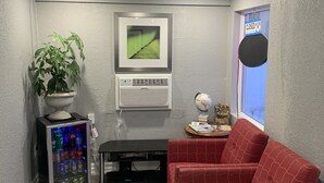A compact living space with a red patterned sofa, a small table with a globe and books, a wall-mounted air conditioner, a beverage cooler, and a framed picture on the wall. - Champlain Motor Inn (Pembroke)