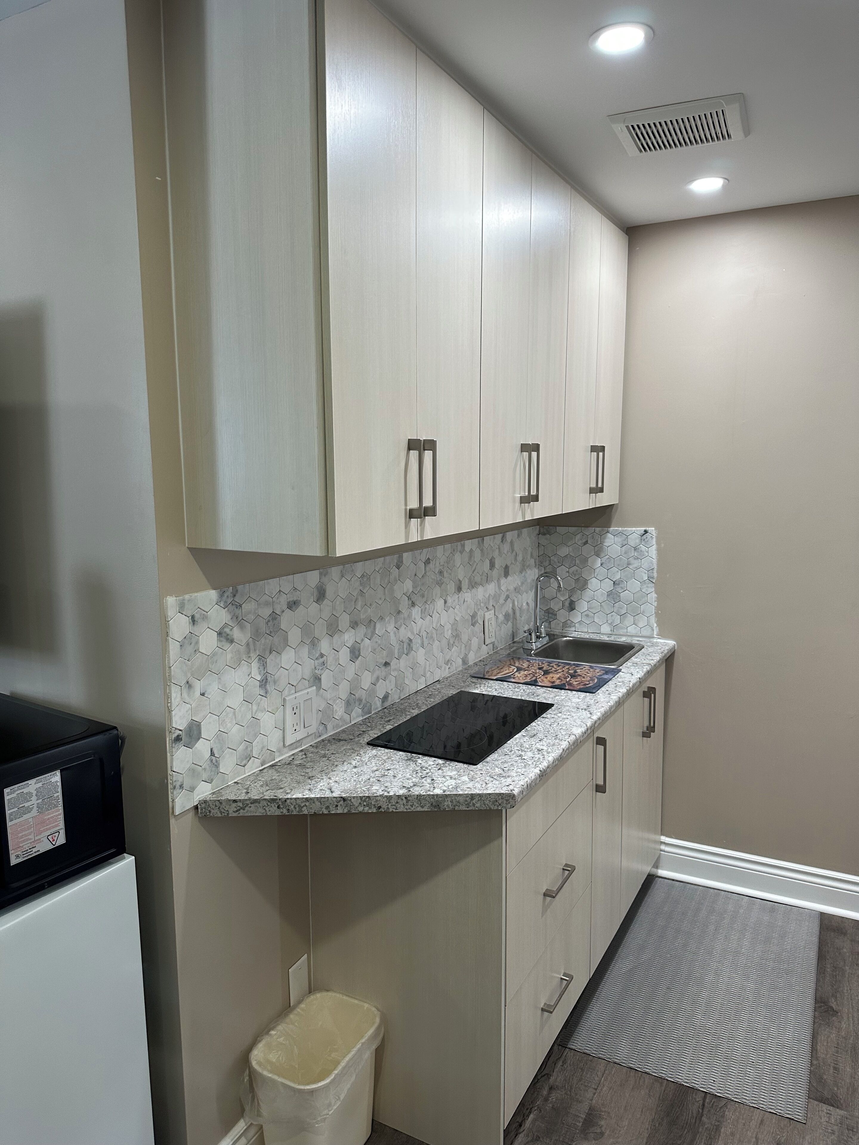 A kitchen with white cabinets, a granite countertop, a hexagonal tile backsplash, a black induction cooktop, a stainless steel sink, and a trash bin.