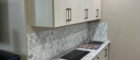A kitchen with white cabinets, a granite countertop, a hexagonal tile backsplash, a black induction cooktop, a stainless steel sink, and a trash bin.