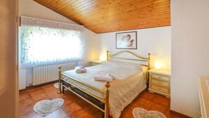 A bedroom with a wooden ceiling, a bed with a gold frame, a bedside table with a lamp, and a window with lace curtains.