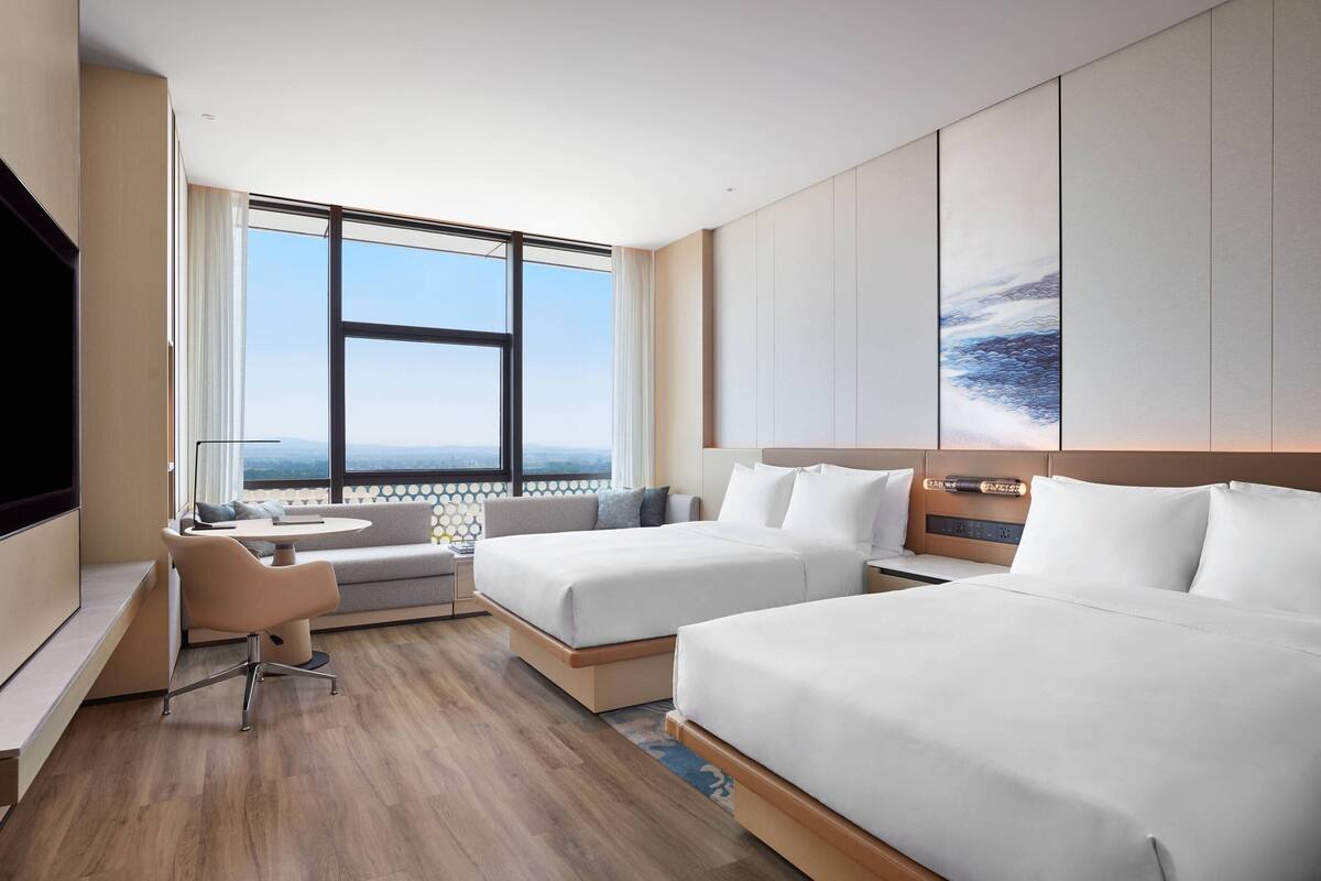 Une chambre d’hôtel moderne équipée de deux lits, d’un canapé, d’un bureau et d’une grande fenêtre donnant sur la ville.