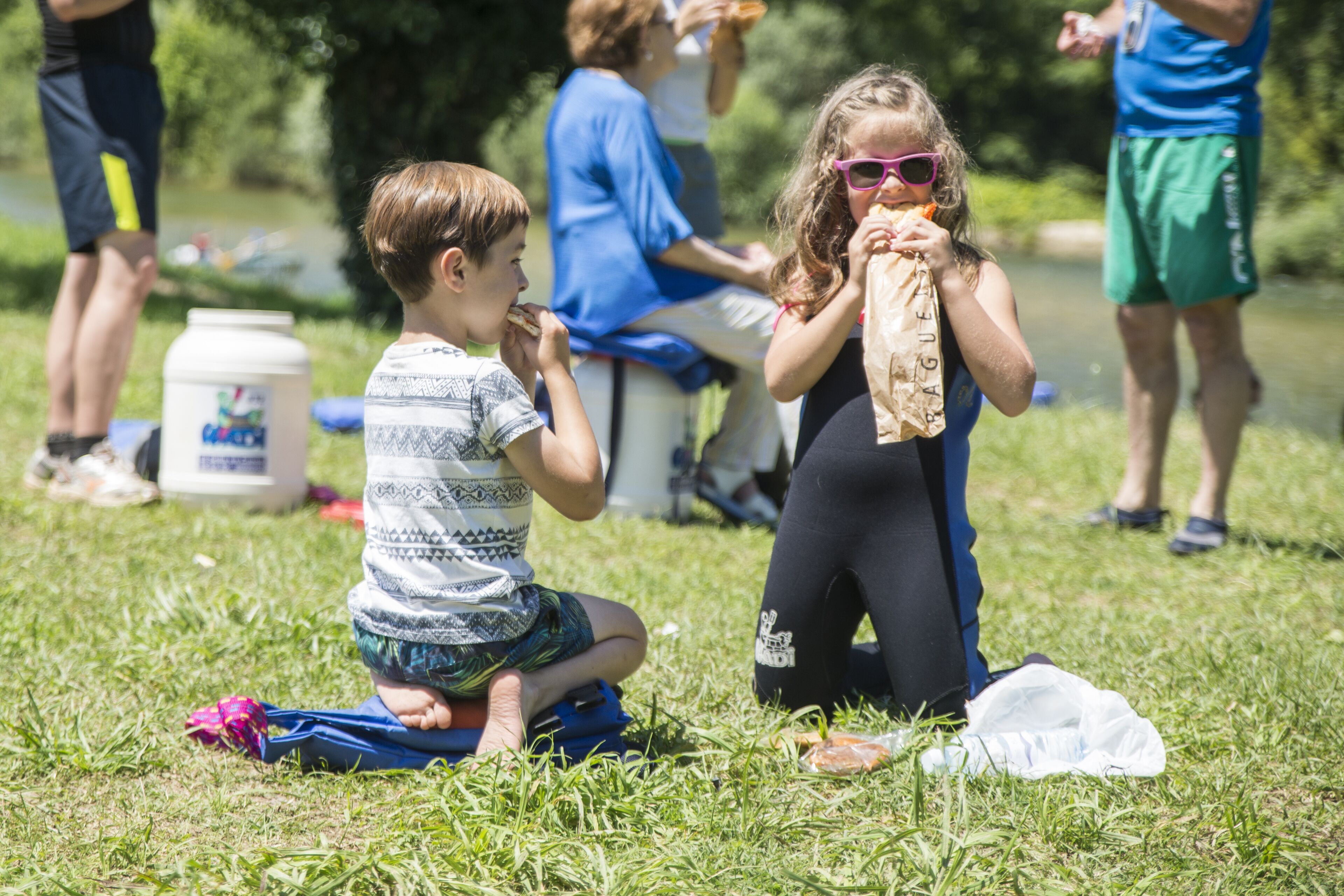 Une zone herbeuse où des personnes sont assises et debout, un plan d’eau en arrière-plan et un conteneur blanc posé sur le sol.