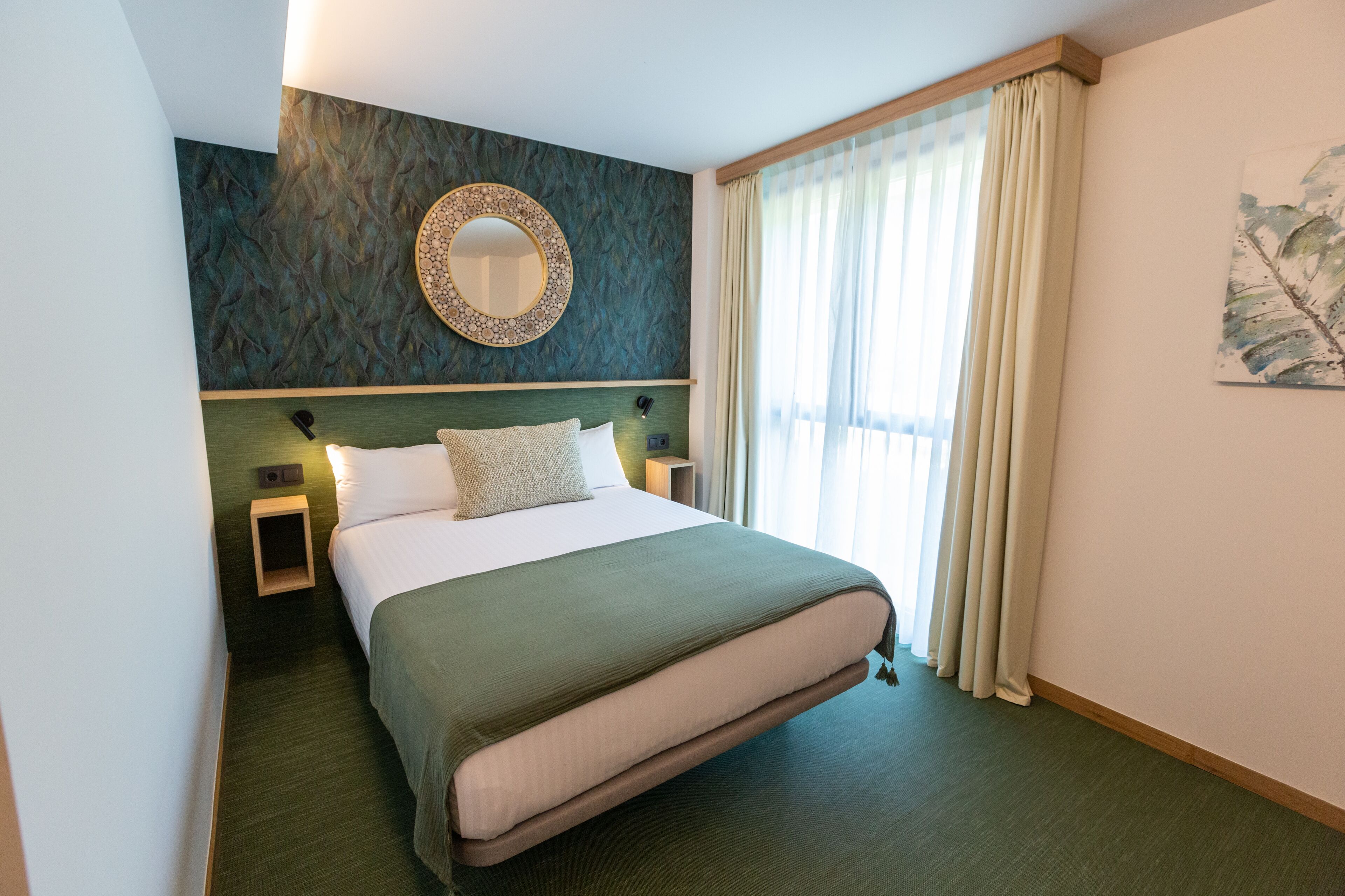 A hotel room with a large bed, a round mirror on the wall, a nightstand, and a window with curtains.