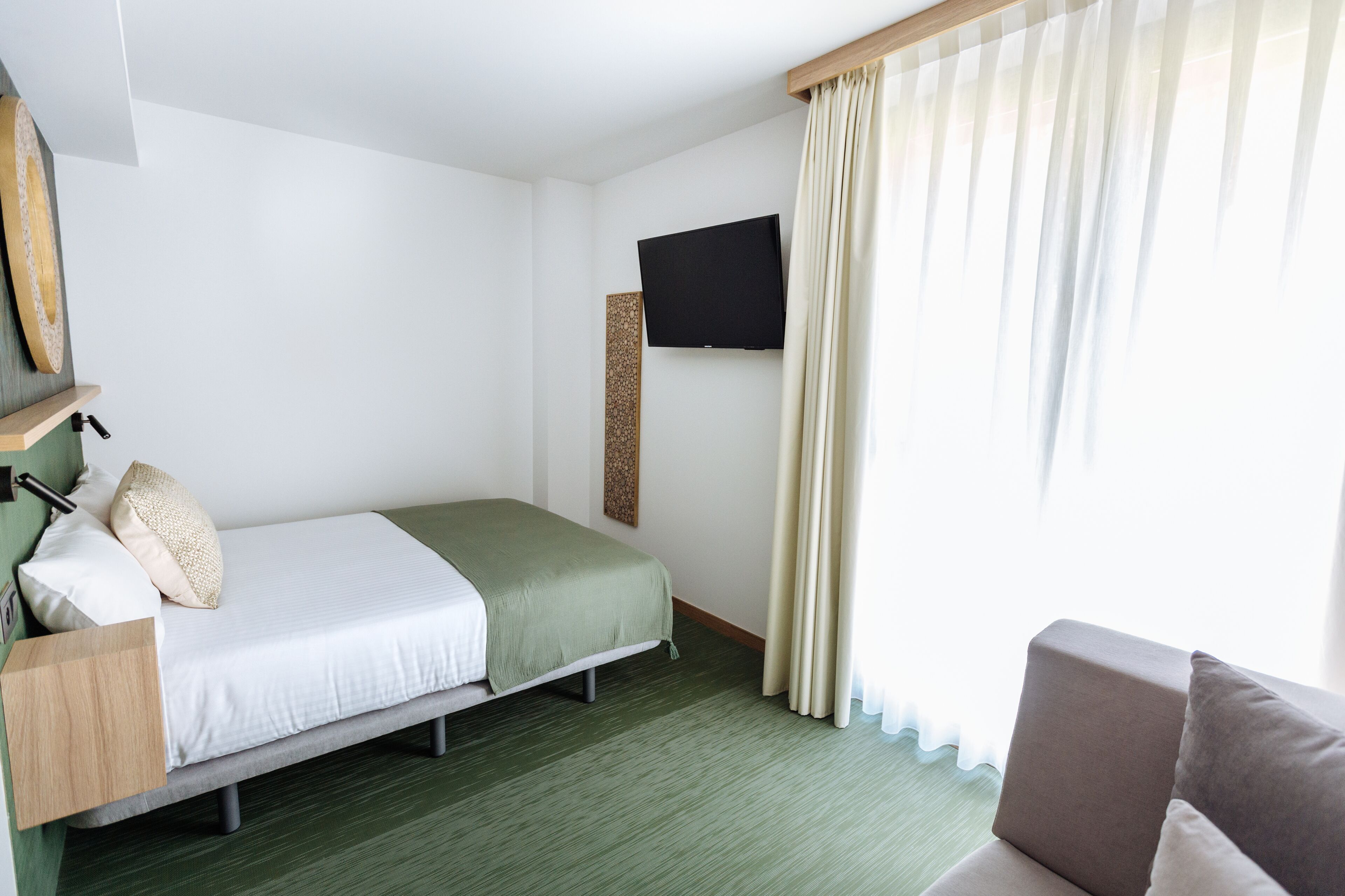 Una habitación de hotel con una cama, un televisor montado en la pared, un estante de madera y una ventana con cortinas.