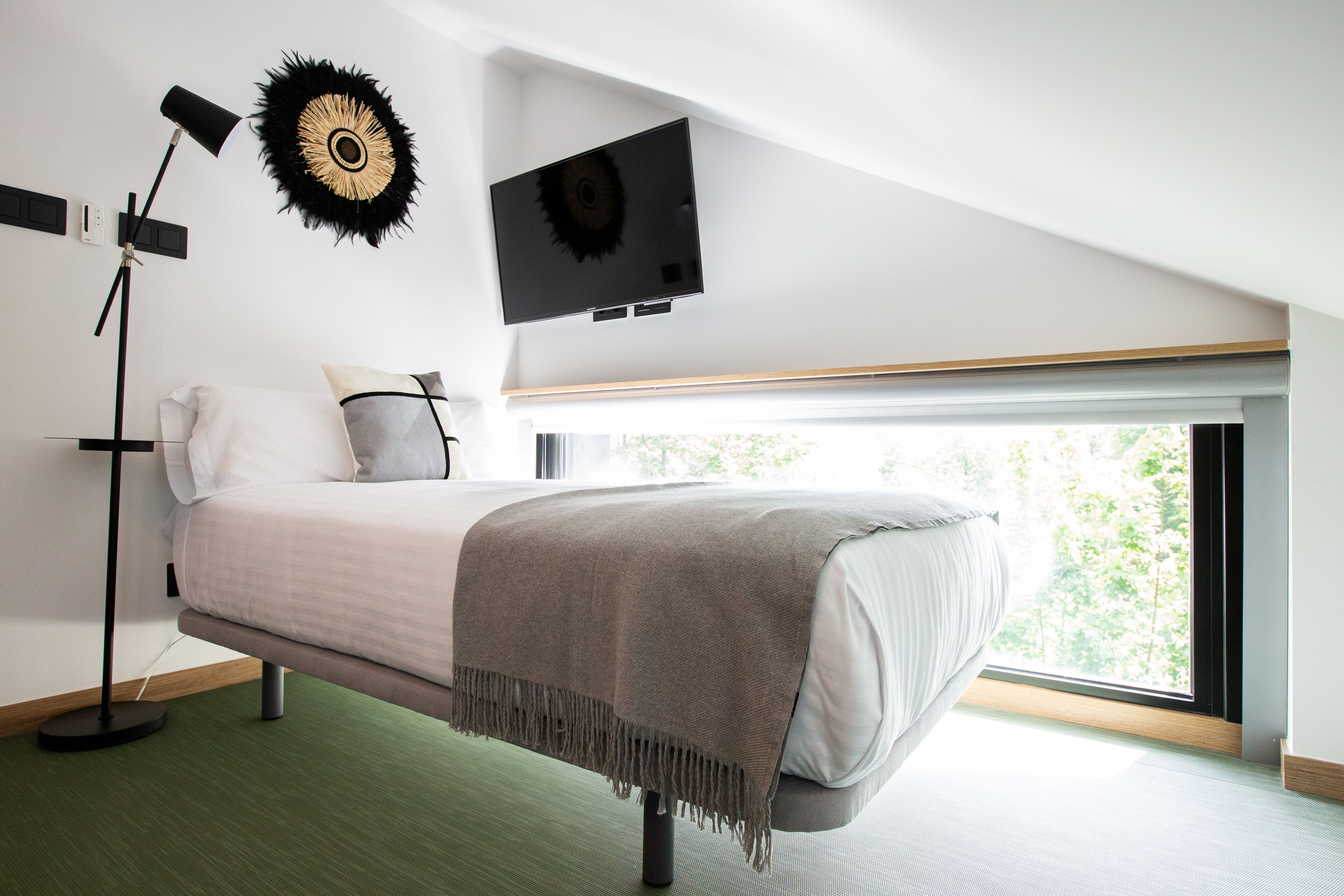 Ein modernes Schlafzimmer mit Bett, Wandfernseher, Stehlampe und großem Fenster mit Blick auf Grünflächen.