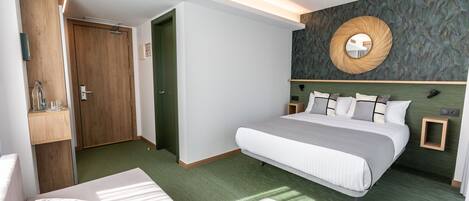A modern hotel room with a large bed, a small seating area, a wooden wardrobe, and a mirror on the wall.