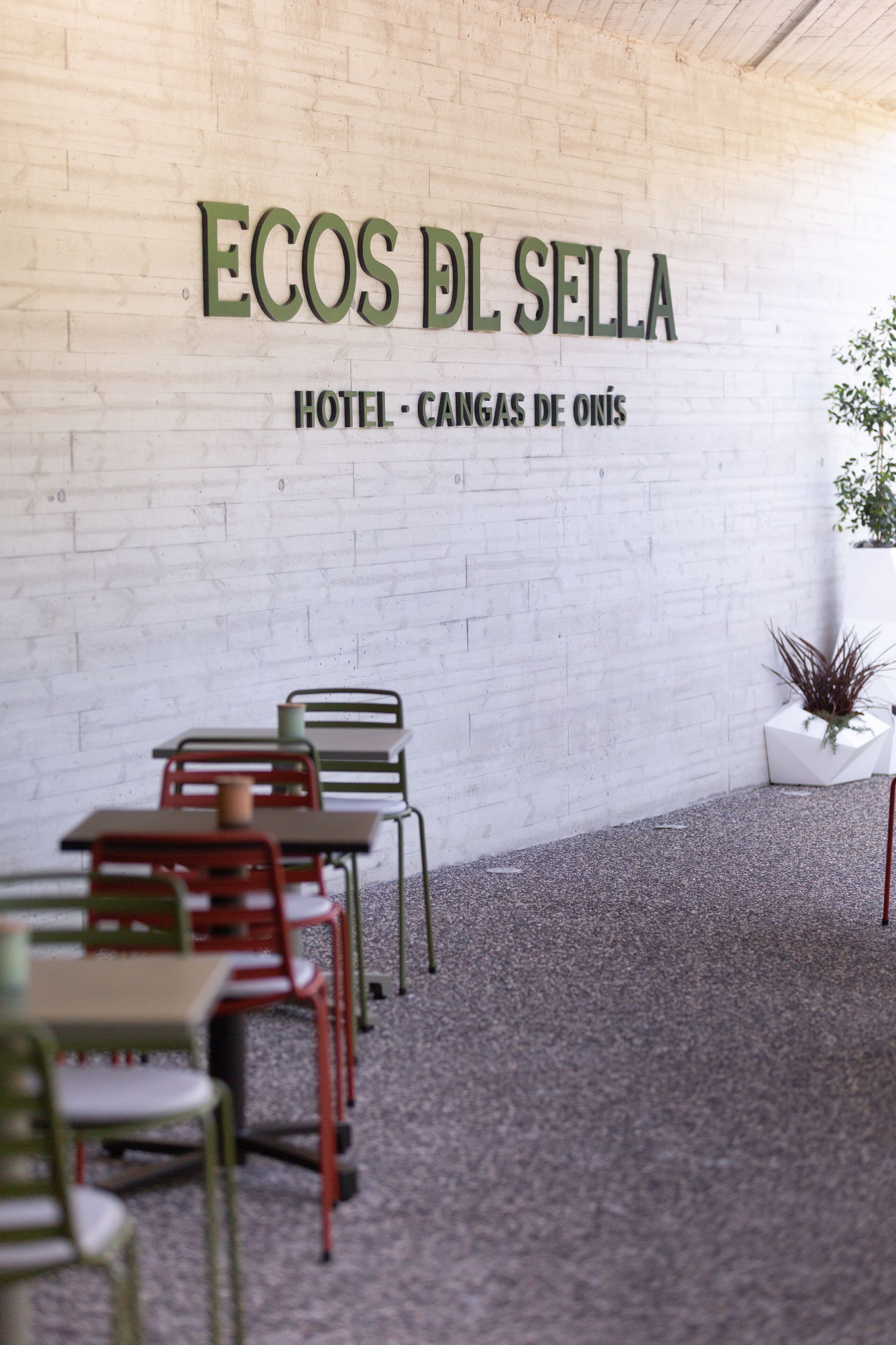 Interior de un hotel con una pared de ladrillo blanco, señalización verde que dice "ECOS DEL SELLA HOTEL - CARGAS DE ONÍS", y varias mesas y sillas.