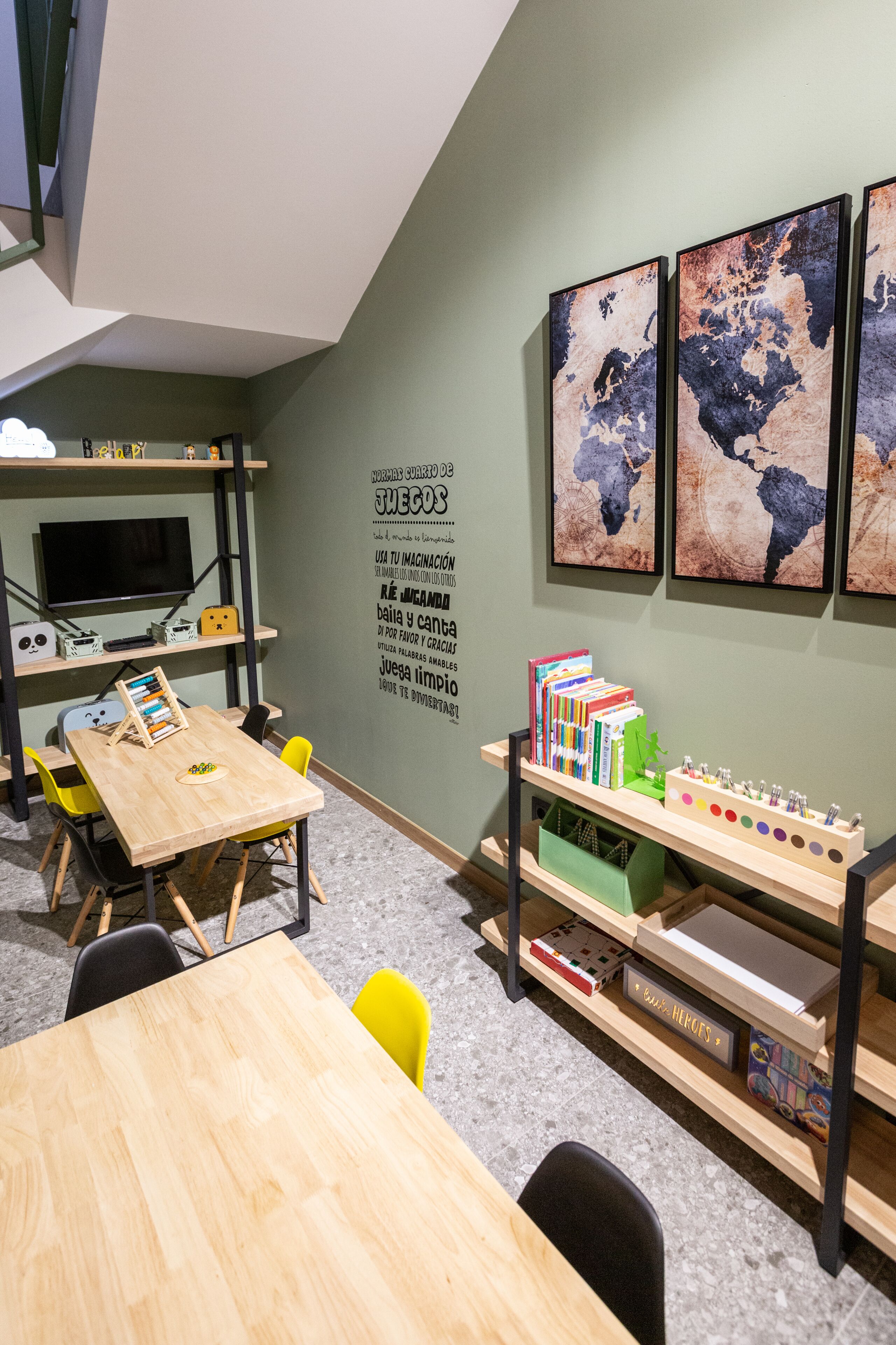 Una habitación moderna y luminosa, con una mesa de madera, sillas amarillas, una estantería llena de libros y juguetes, un televisor de pantalla plana y un cuadro con un mapa del mundo en la pared.