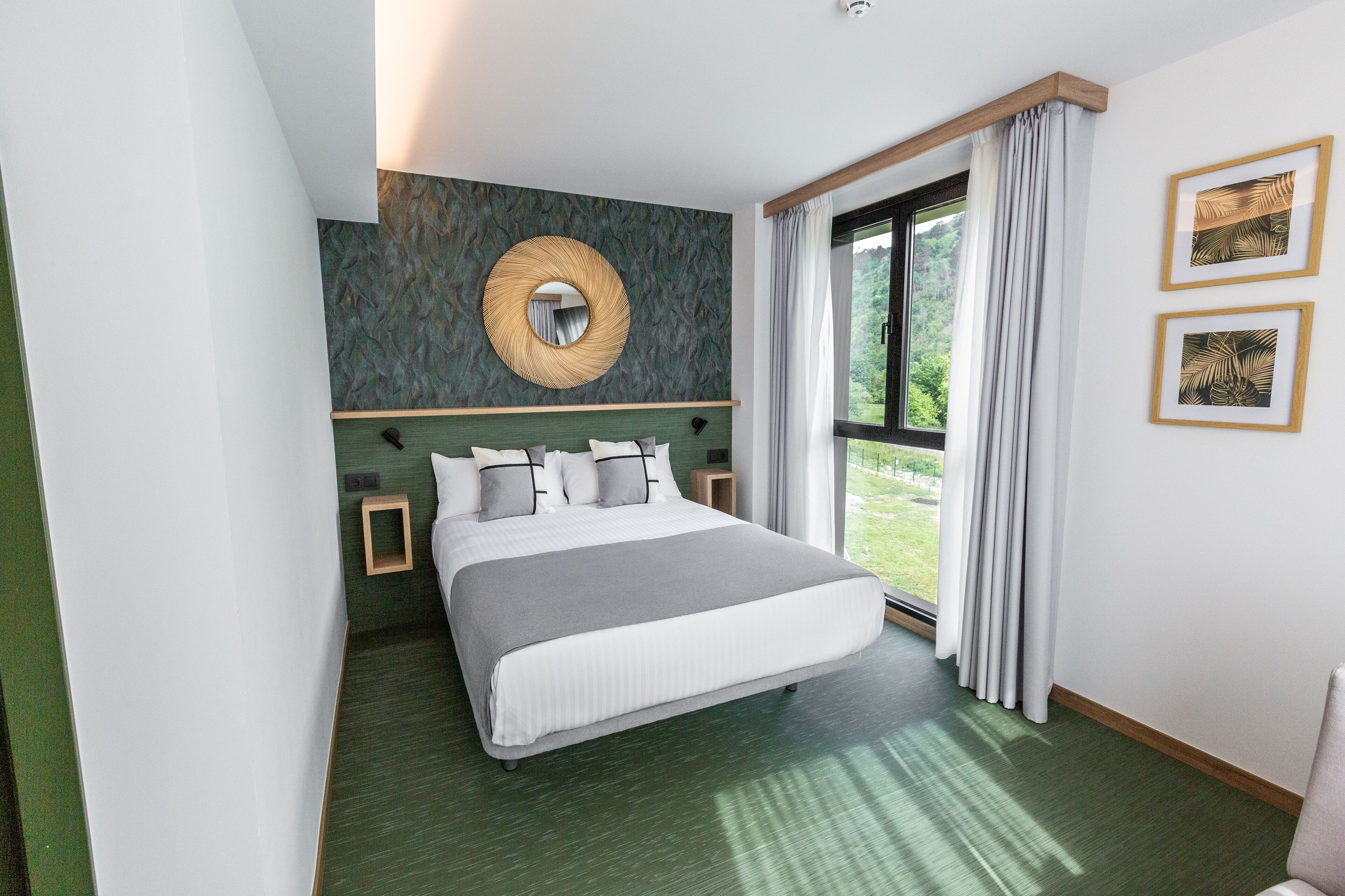 A modern hotel room with a large bed, a round mirror, a view of greenery, and two framed pictures on the wall.