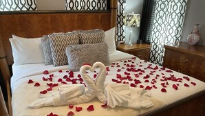 A neatly made bed with rose petals and two swan-shaped towels, a patterned pillow, a bedside lamp, and a decorative vase on a wooden nightstand. - Peace & Plenty Inn Bed & Breakfast (St. Augustine)