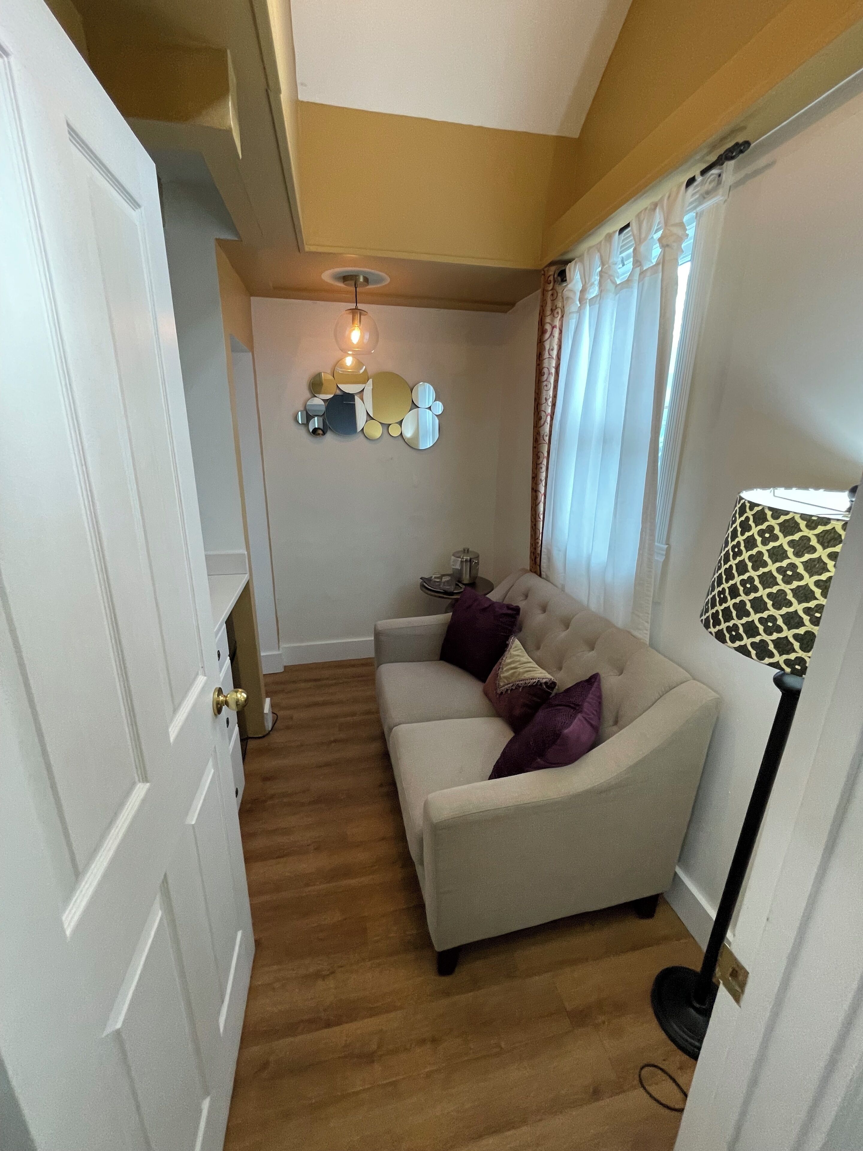 Een kamer met een beige bank, een witte deur, een plafondlamp en een lamp met een patroon.