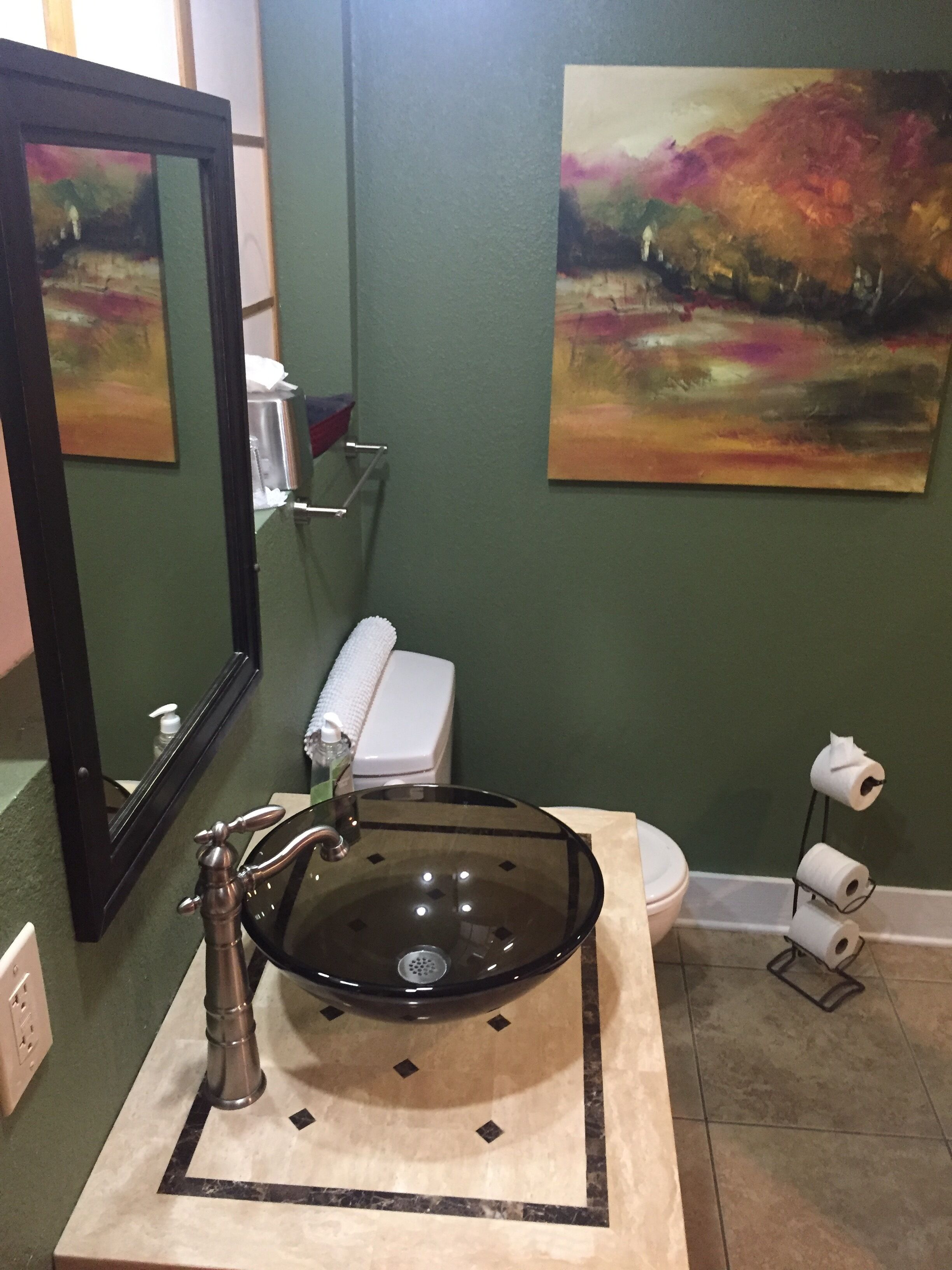 A bathroom with a glass sink, a toilet, and two paintings on the wall.