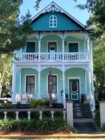Una casa de dos pisos con fachada verde y detalles blancos, que incluye un balcón con barandillas blancas y una puerta principal con un ventanal decorativo.