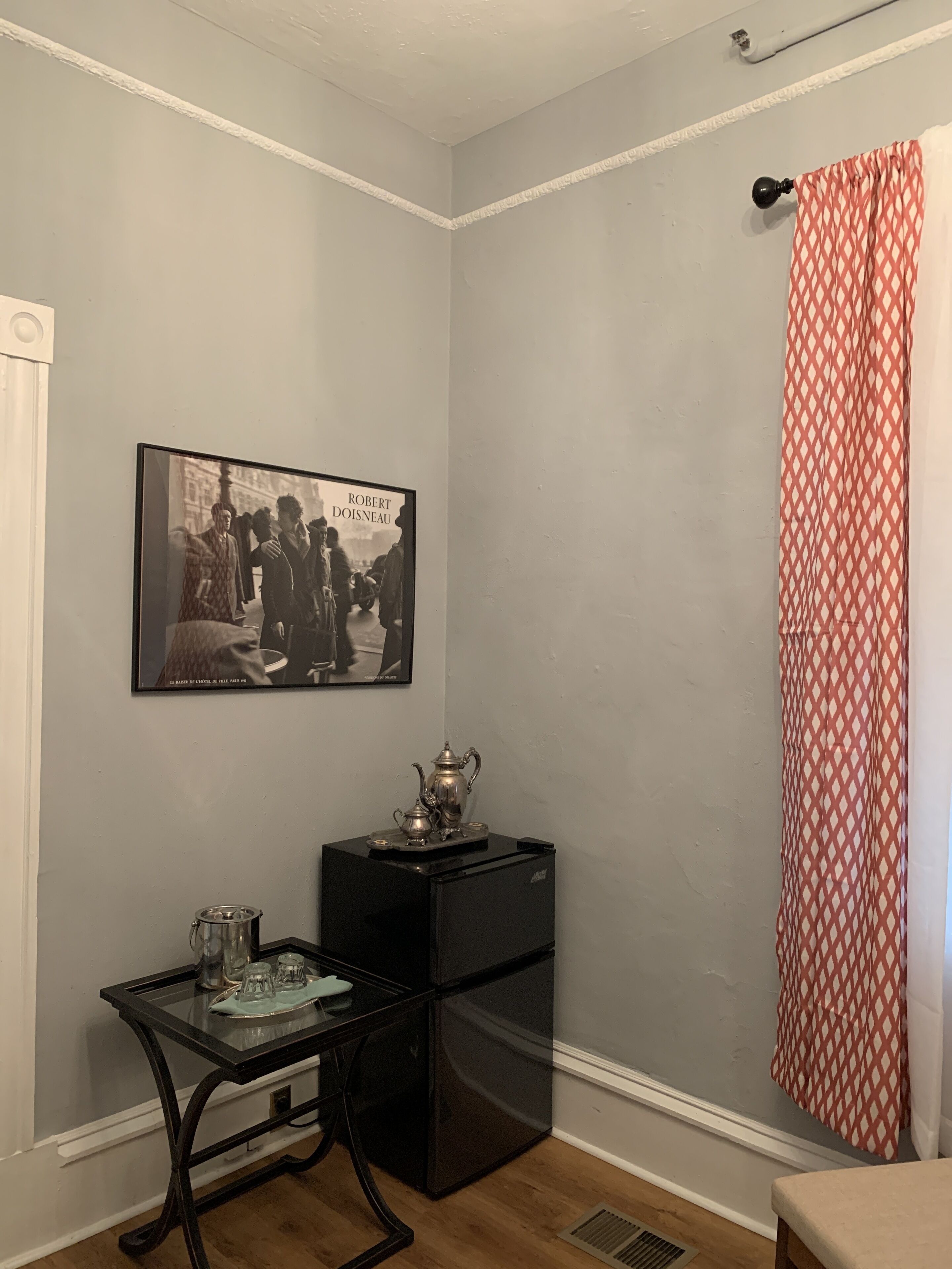 A corner room with a black refrigerator, a glass table with a tray, a framed black and white photo, and a red patterned shower curtain.