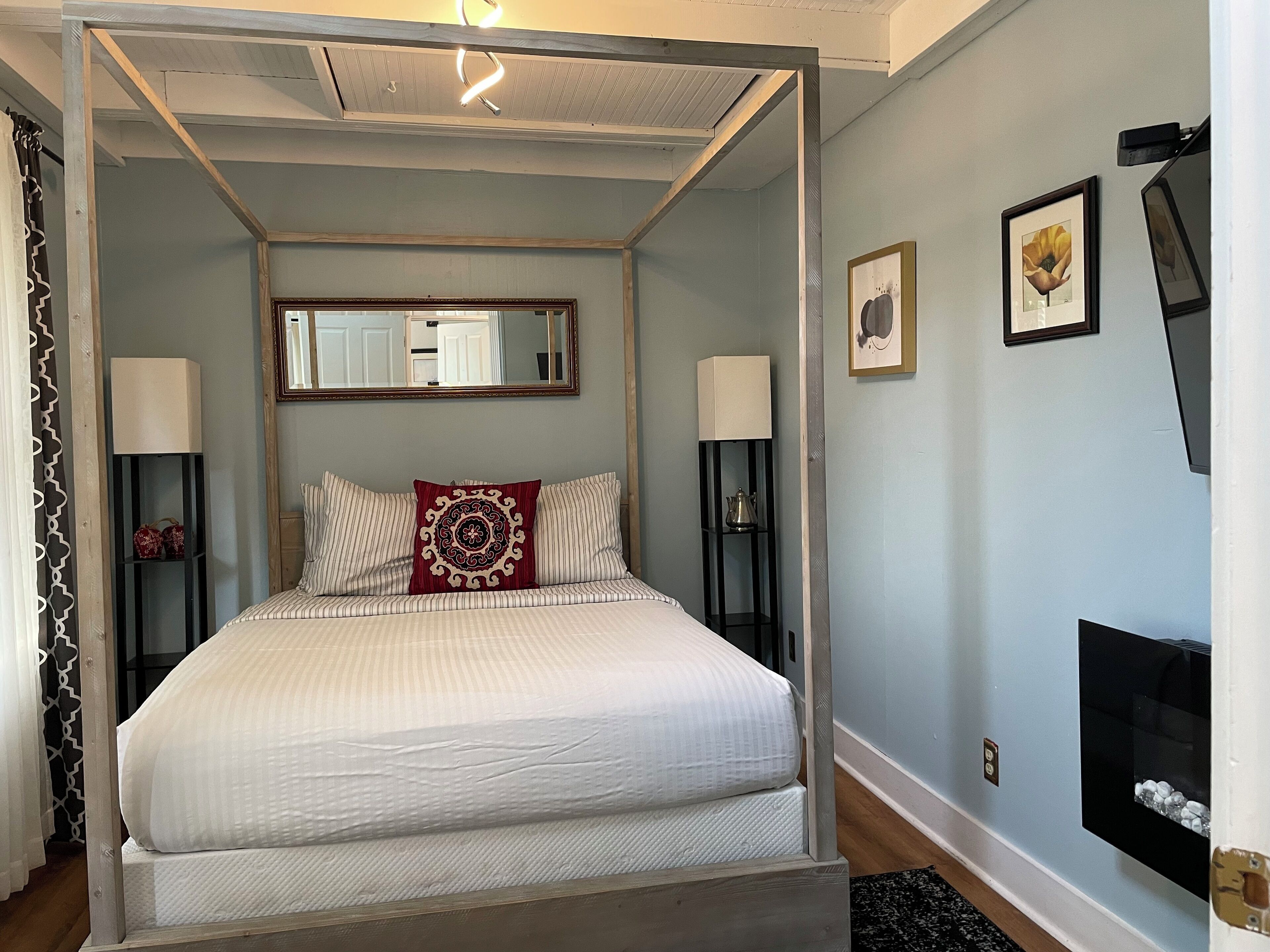 Un lit à baldaquin avec une couvre-lit blanche, deux oreillers et un coussin décoratif rouge. Il y a deux tables de chevet avec des lampes, un miroir au-dessus du lit et des œuvres d’art encadrées accrochées au mur.