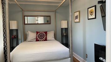 Un lit à baldaquin avec une couvre-lit blanche, deux oreillers et un coussin décoratif rouge. Il y a deux tables de chevet avec des lampes, un miroir au-dessus du lit et des œuvres d’art encadrées accrochées au mur.