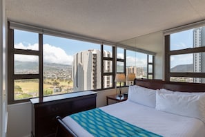 A hotel room with a large bed, a nightstand, a lamp, and a view of a cityscape through large windows.