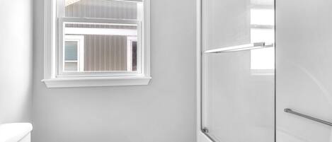 A bathroom with a toilet, a shower with a glass door, and a window with blinds.