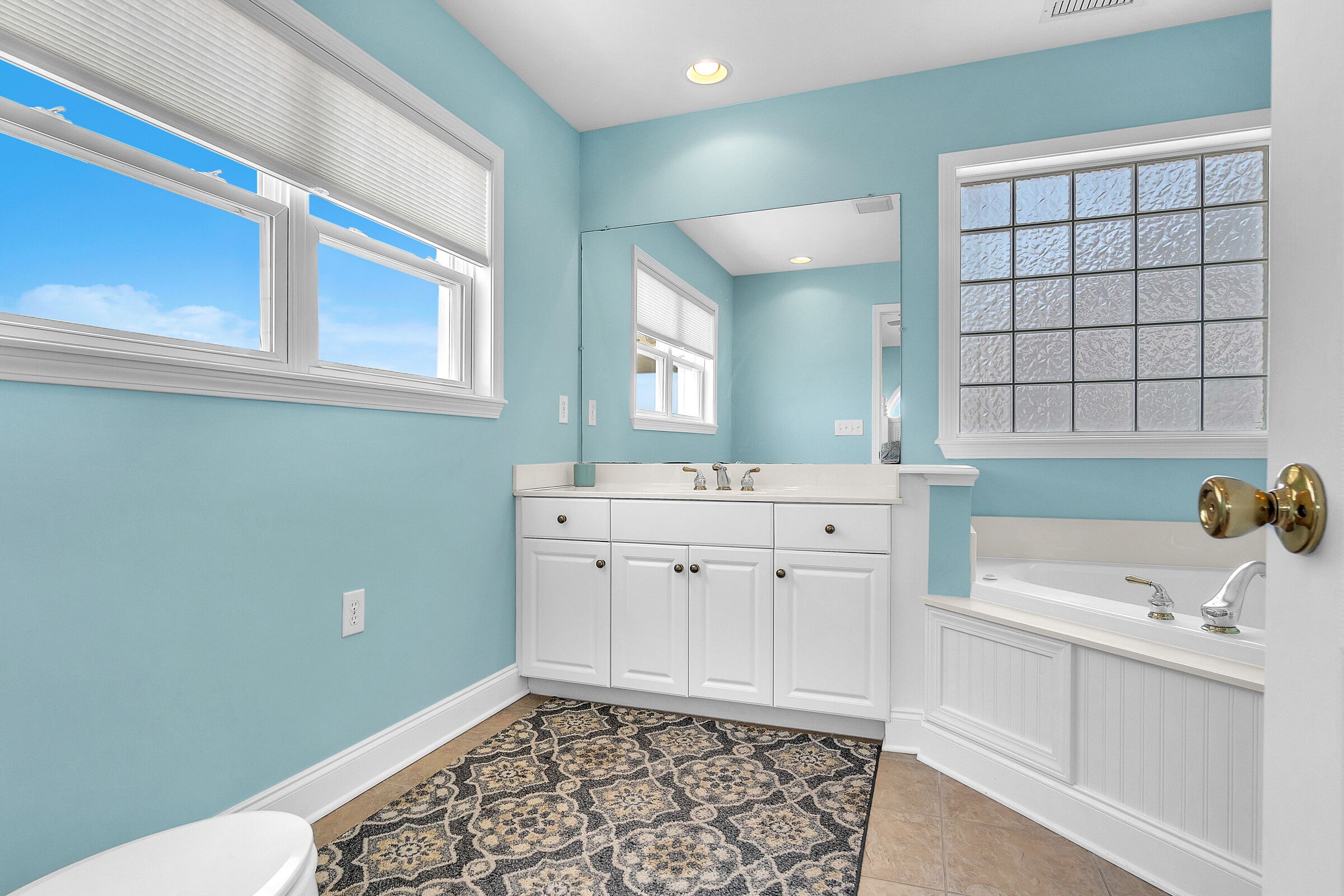 A bathroom with a blue wall, white cabinets, a patterned rug, and a large window with blinds.