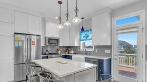 Een moderne keuken met roestvrijstalen apparatuur, een groot kookeiland met barkrukken, hanglampen en uitzicht door een glazen deur.