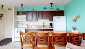 Una cocina con armarios de madera, un refrigerador blanco, un microondas, una estufa y un fregadero. Hay tres sillas de madera con asientos acolchados.