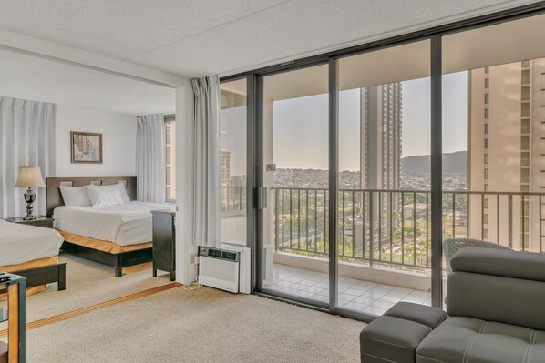A hotel room with a bed, bedside table, lamp, air conditioning unit, and a view of the city through large windows. - Excellent Diamond Head View Condo - Remodeled, Free Parking! (Honolulu)