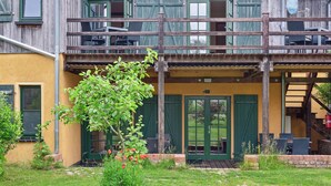 Un edificio rustico con facciata in legno, un balcone con sedie e un piccolo giardino con un albero e fiori.