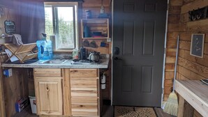 A compact kitchen with wooden cabinets, a sink, and a stove. There is a window providing natural light and a door to the right. - Rustic Moose Cabin Getaway - Glamping (Cantwell)