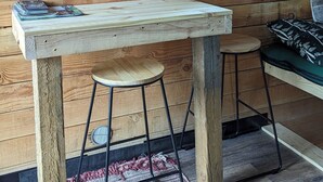 A wooden cabin interior with a wooden table, two stools, a window with curtains, and a painting on the wall. - Rustic Moose Cabin Getaway - Glamping (Cantwell)
