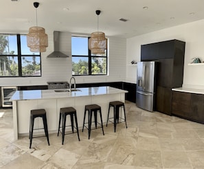 A modern kitchen with a central island, bar stools, a stainless steel refrigerator, and large windows.